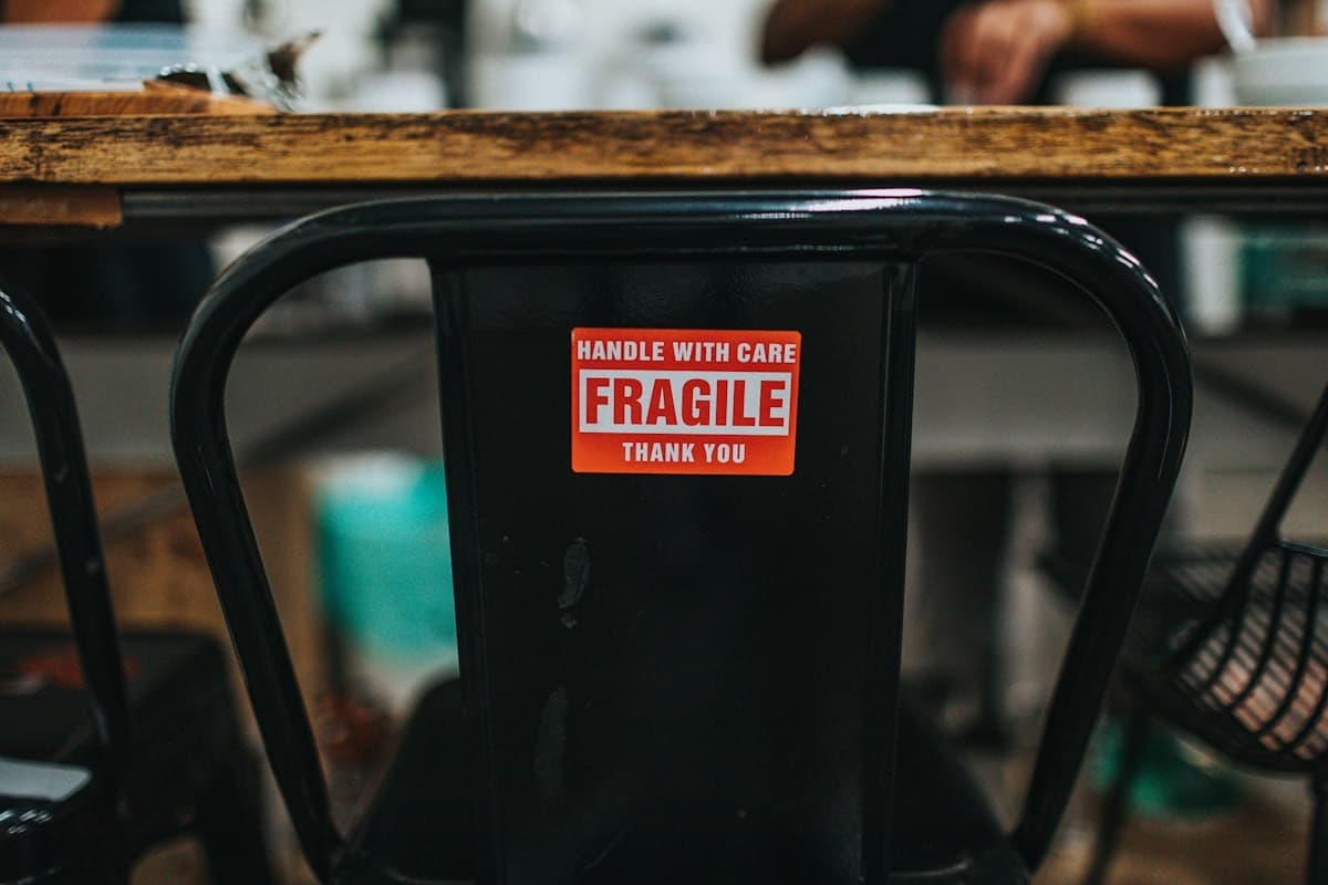 Raudona lipni įspėjamoji etiketė „Fragile — handle with care“, priklijuota ant metalinės kėdės atlošo