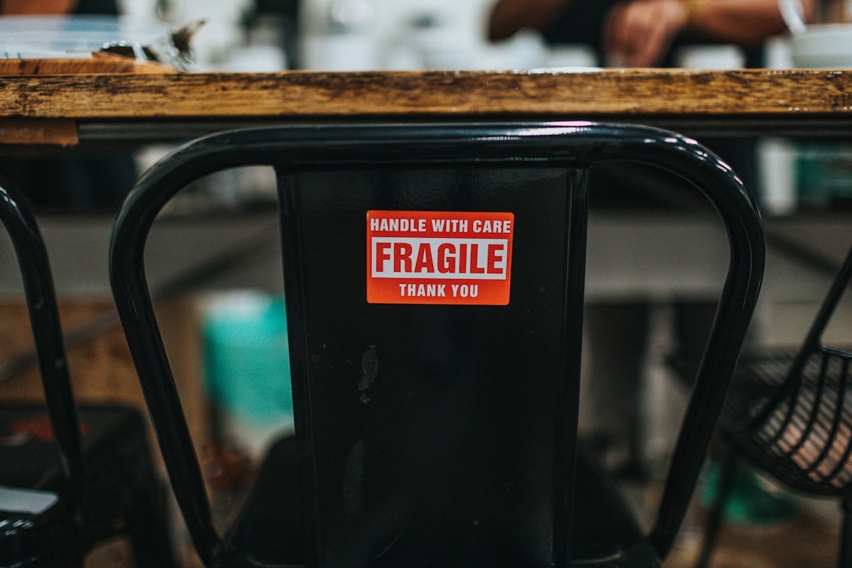 Raudona lipni įspėjamoji etiketė „Fragile — handle with care“, priklijuota ant metalinės kėdės atlošo